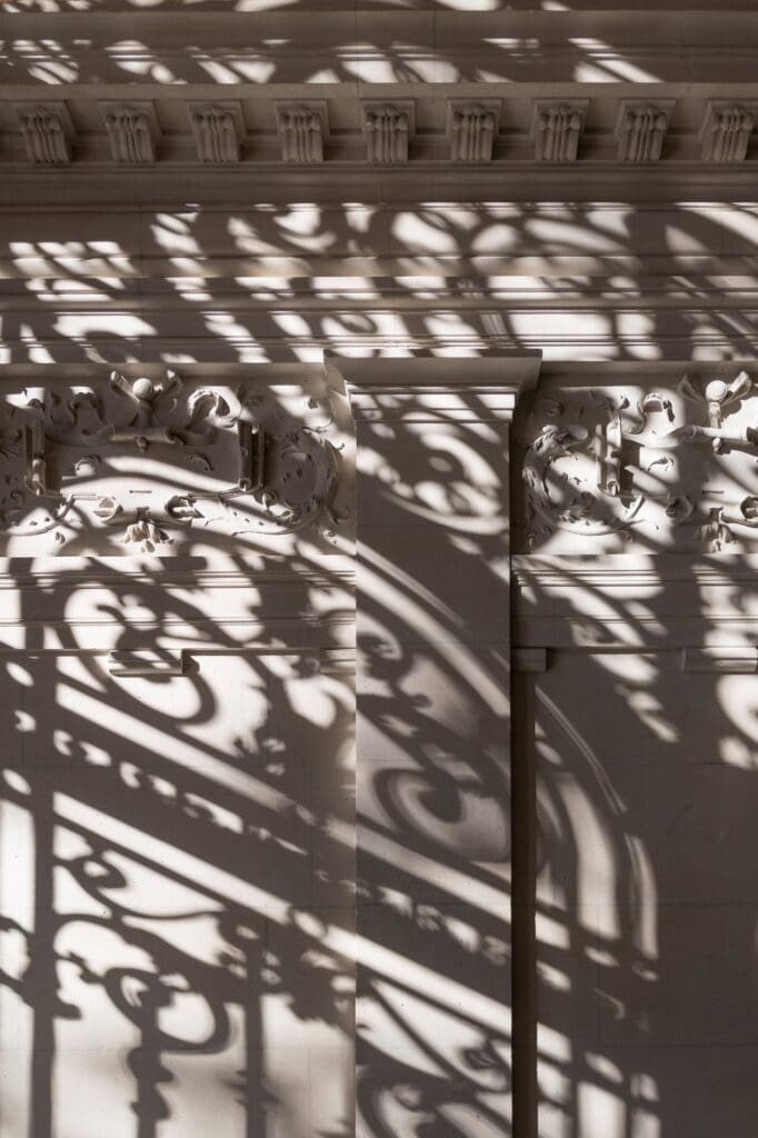 Shadows of ornate wrought ironwork falling across carved stone ornament and a dentil cornice at Apple Champs-Élysées