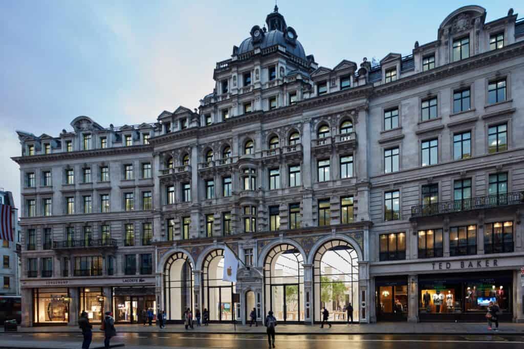 Apple Regent Street in London at dusk, the domed 1898 Portland stone building with four tall glazed arches lit from within, flanked by its 1920s wings and shoppers on the pavement