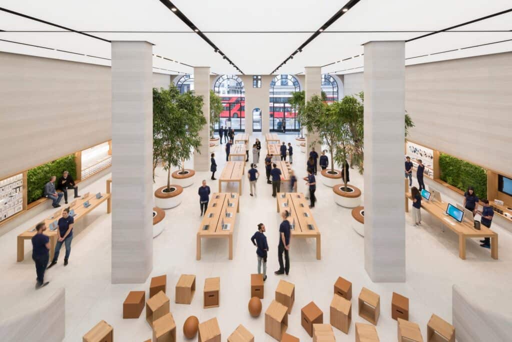 Grand hall of Apple Regent Street seen from the mezzanine, oak tables and trees between stone-clad columns under the luminous ceiling, Forum cubes in front and red buses in the arched windows