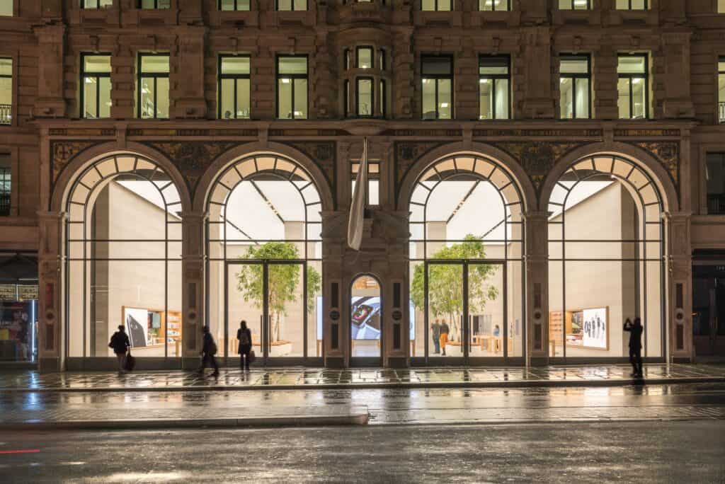 Night view of Apple Regent Street from the wet street, four full-height arched windows framing the trees and glowing interior beneath the stone facade