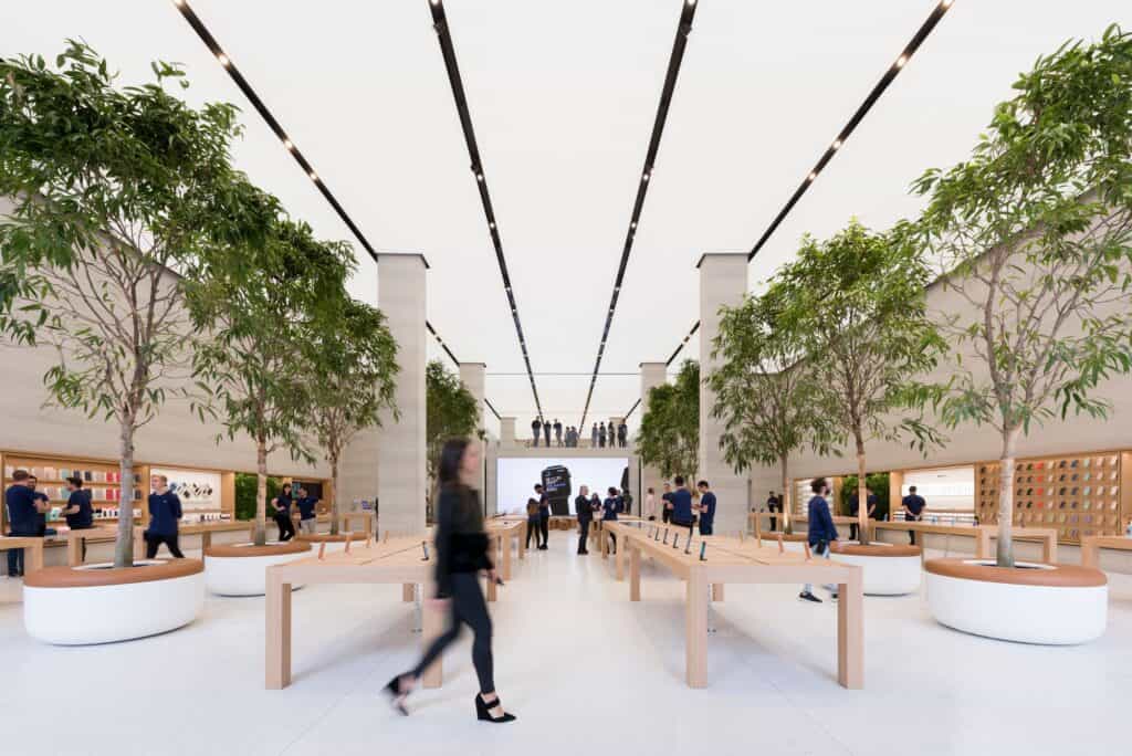 Double-height hall of Apple Regent Street with rows of Ficus trees and oak tables under the long luminous ceiling panels, leading to the video wall of the Forum