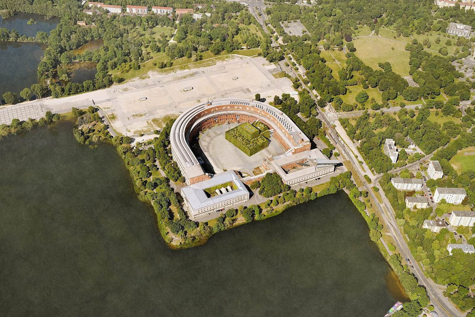 Aerial render of a curved brick congress hall on a lakeside, surrounded by trees, open paving and water