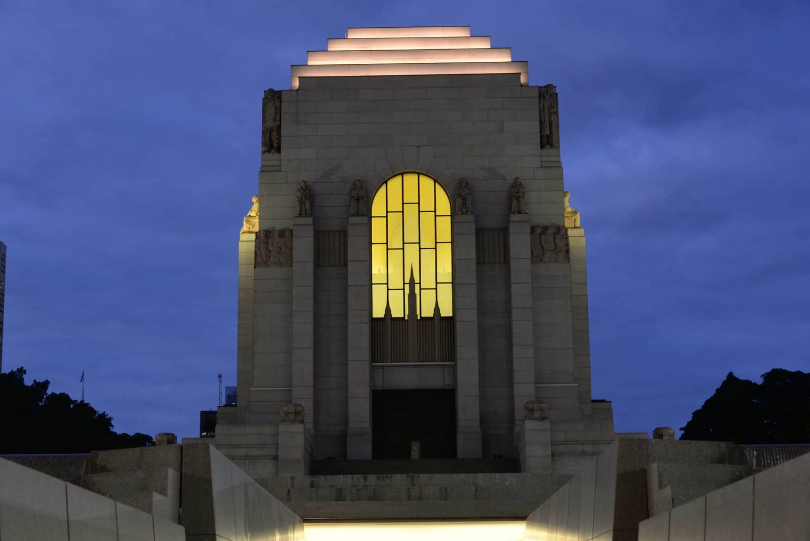 The Anzac Memorial in Hyde Park, Sydney, at dusk, with its tall amber window glowing, a stepped top and sculpted figures on the buttresses