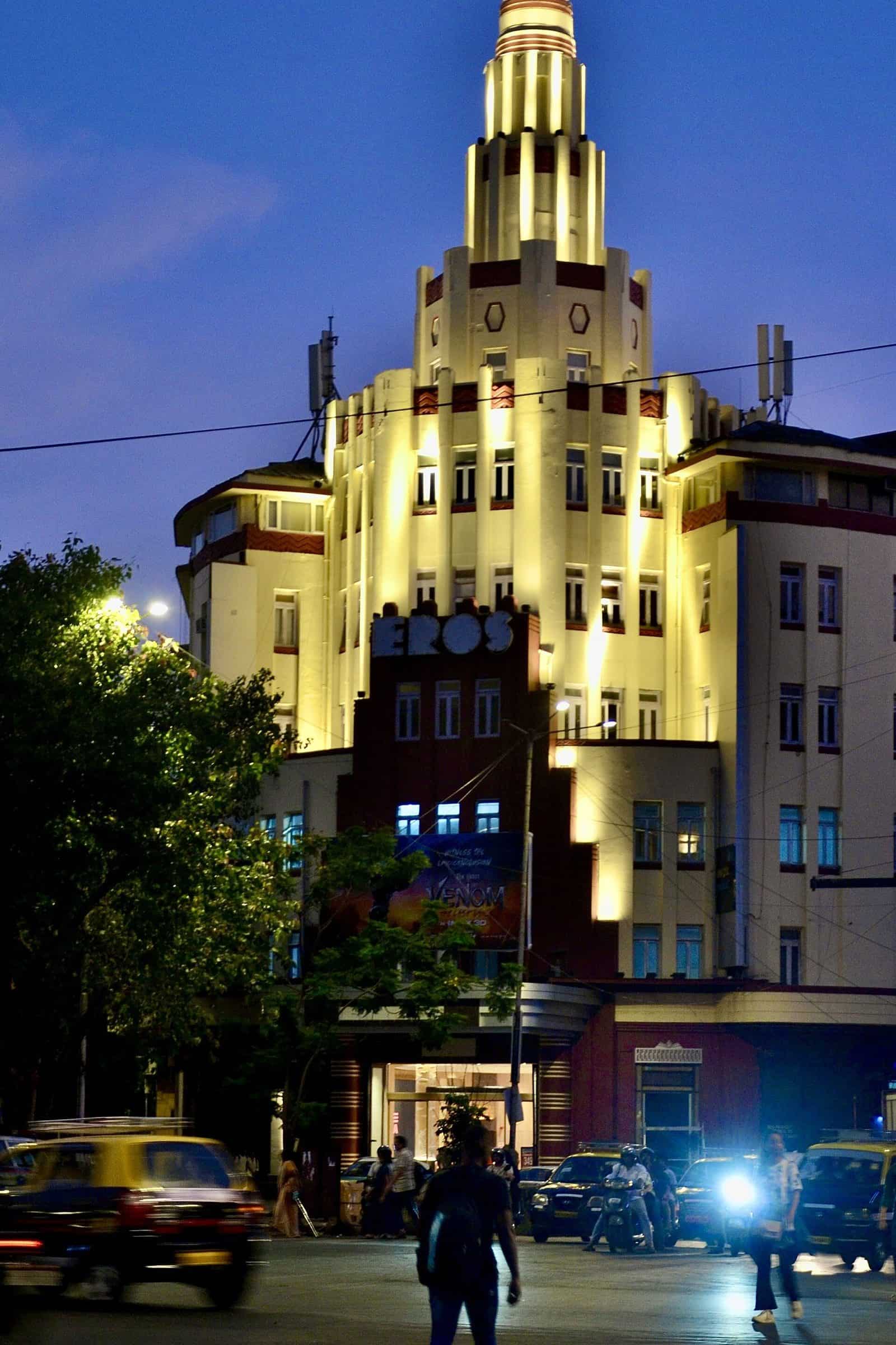 The floodlit cream and red tower of the Eros Cinema in Mumbai at dusk, with stepped tiers rising to a rounded top and street traffic and pedestrians below