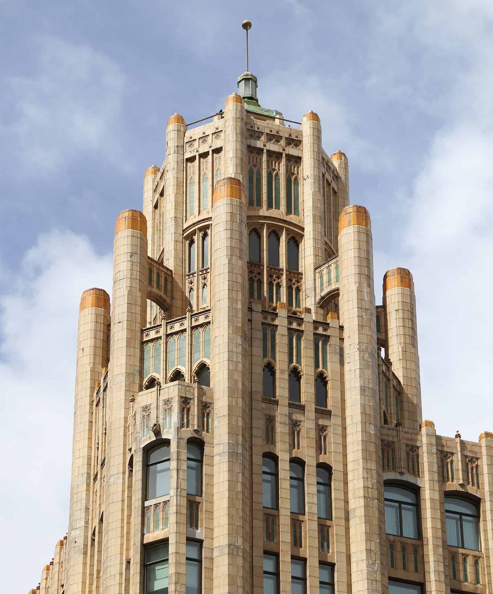 The cream terracotta tower of the Grace Building in Sydney, with rounded vertical piers topped in gold, Gothic-style window tracery and a small green cupola against a cloudy sky