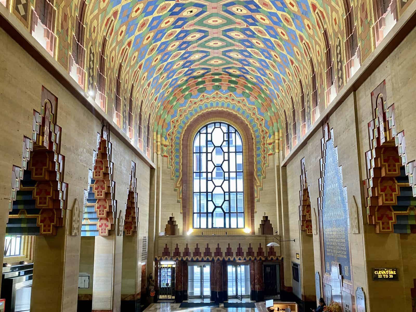The vaulted lobby of the Guardian Building in Detroit, with a ceiling of colourful geometric tiles, stepped orange and red patterns on the walls and a tall leaded window at the end