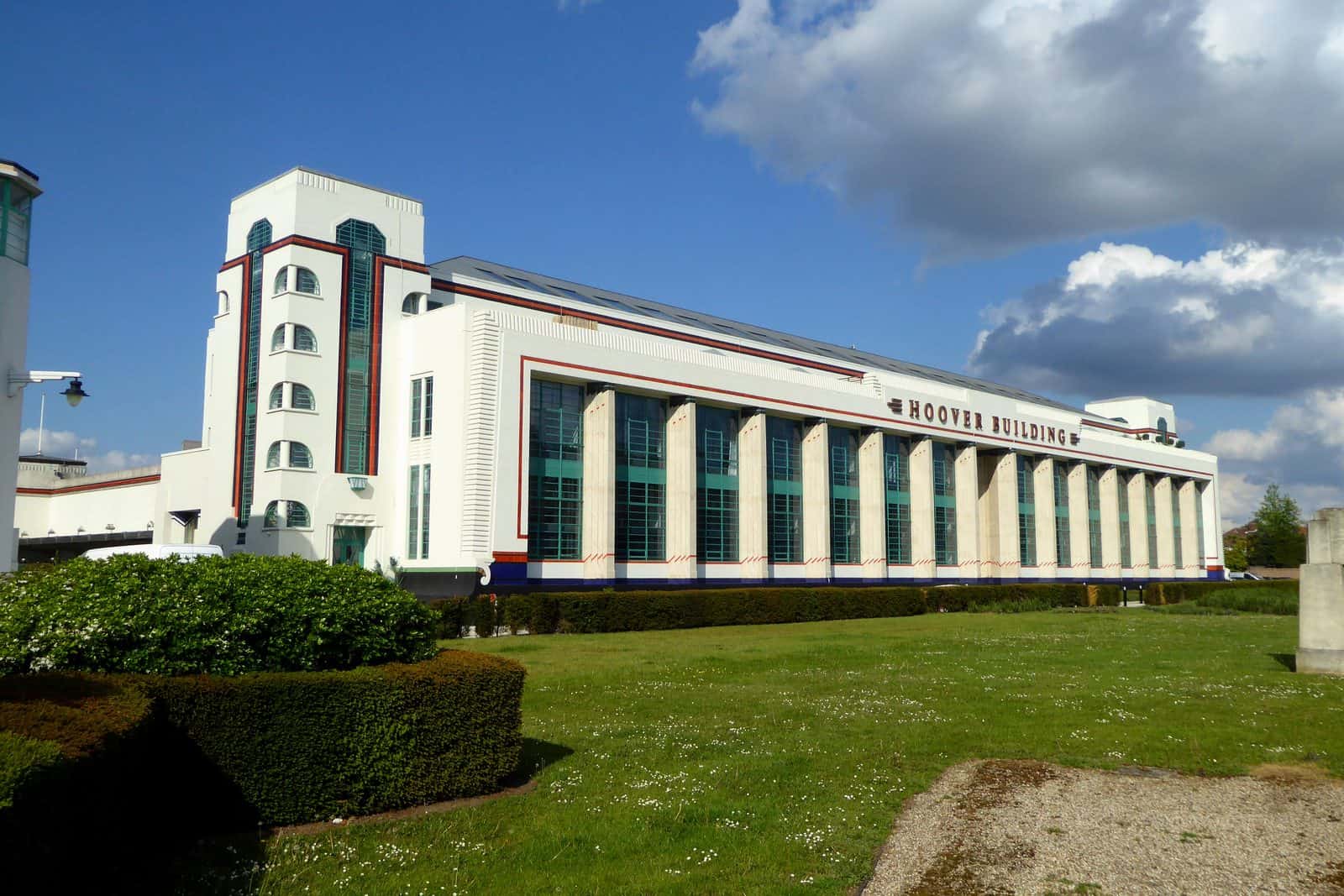 The long white facade of the Hoover Building in Perivale, west London, with a corner stair tower, green glazing, red trim and Hoover Building lettering, behind a lawn and hedges