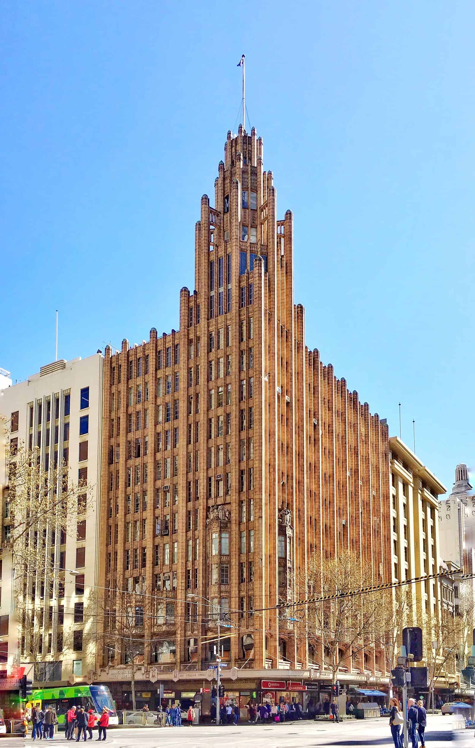 The Manchester Unity Building in Melbourne, a terracotta-clad corner tower with ribbed vertical piers stepping up to a spire, seen from the corner of Collins and Swanston Streets with a tram below