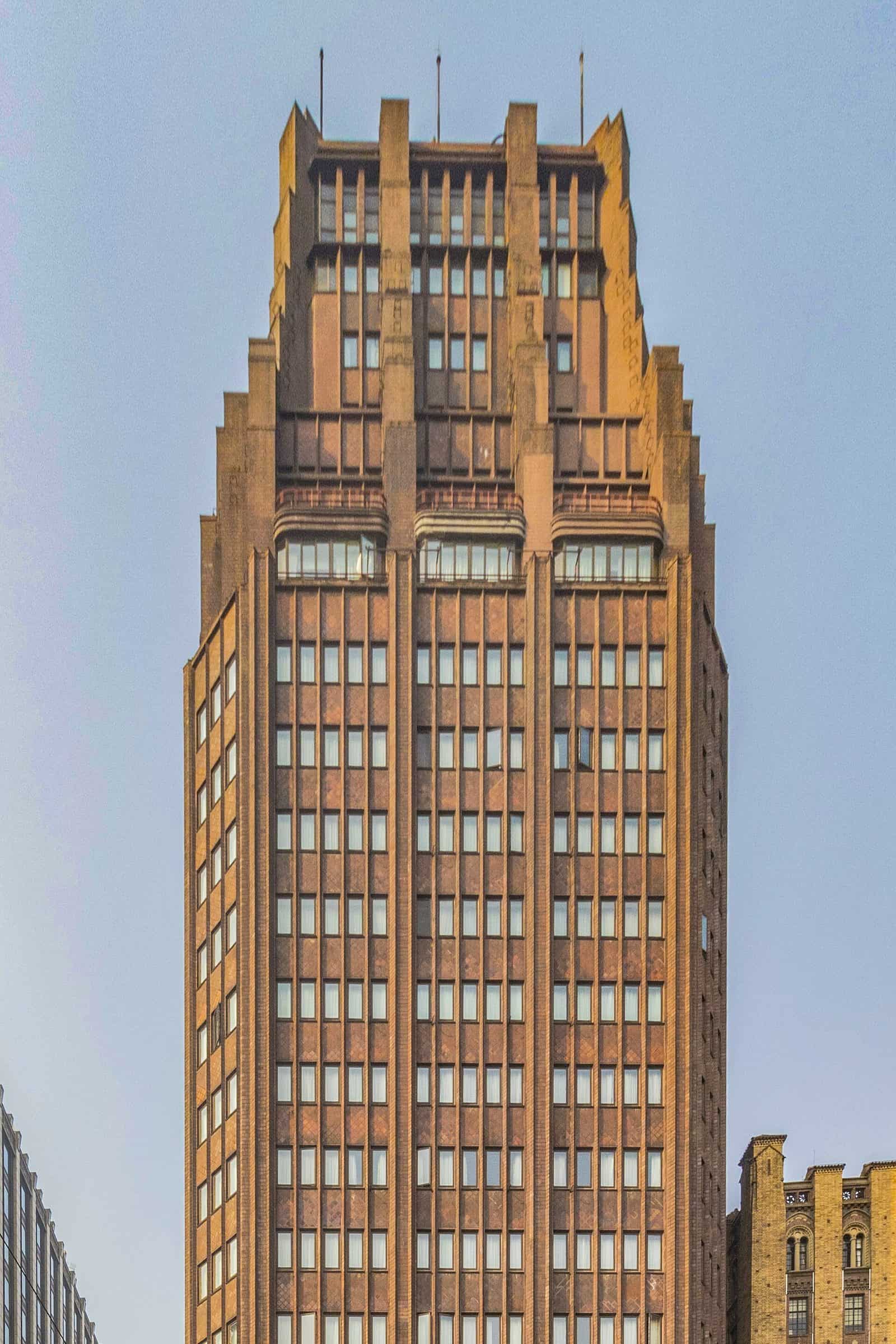 The dark brown brick tower of the Park Hotel in Shanghai, with strong vertical piers and a stepped top rising against a pale sky