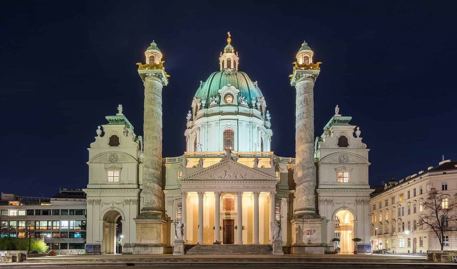 The Karlskirche in Vienna lit up at dusk, with its green oval dome, a temple portico and two tall carved columns either side against a deep blue sky