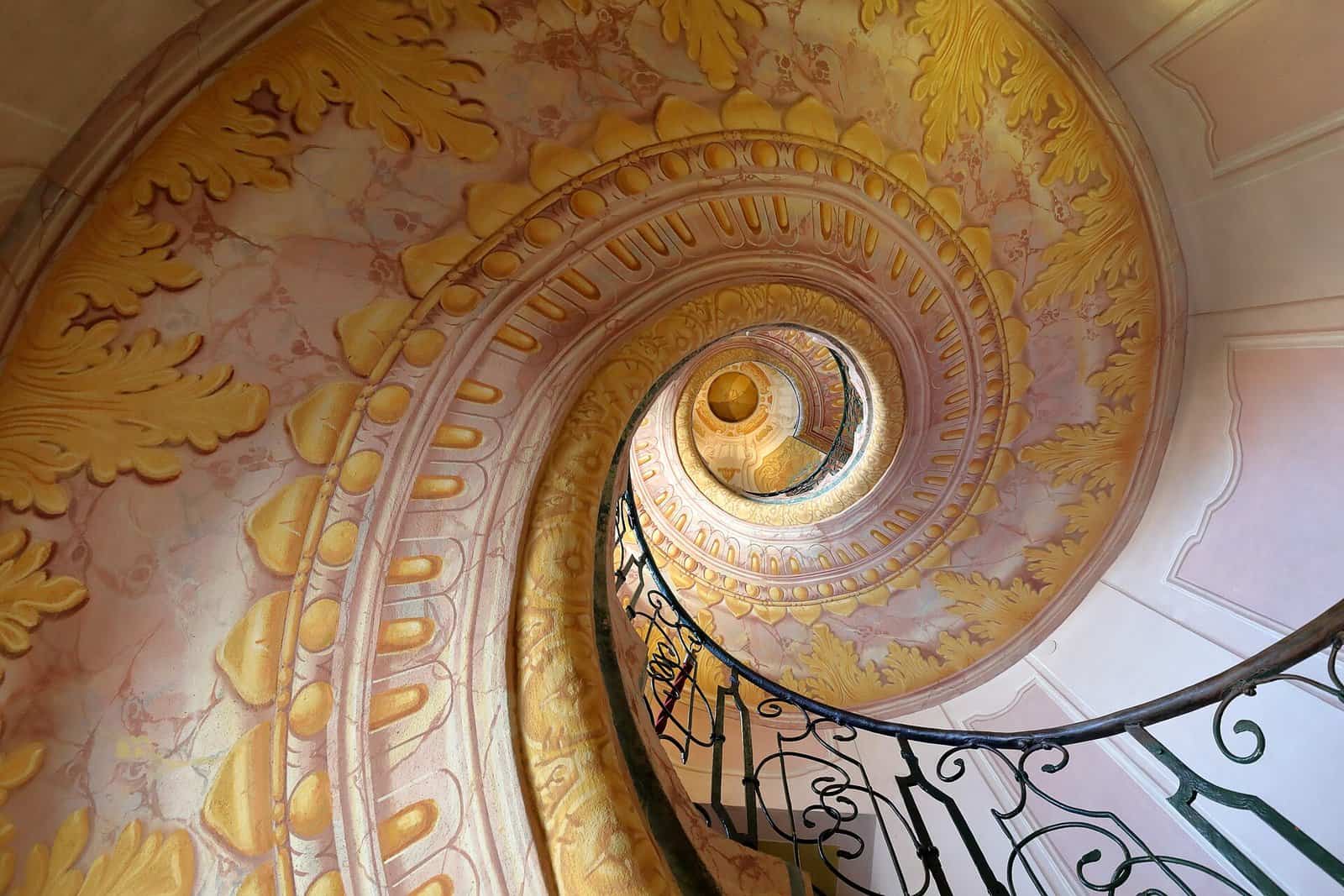 Looking down the curling spiral staircase of Melk Abbey in Austria, with painted pink and gold walls and a wrought-iron balustrade winding into the centre