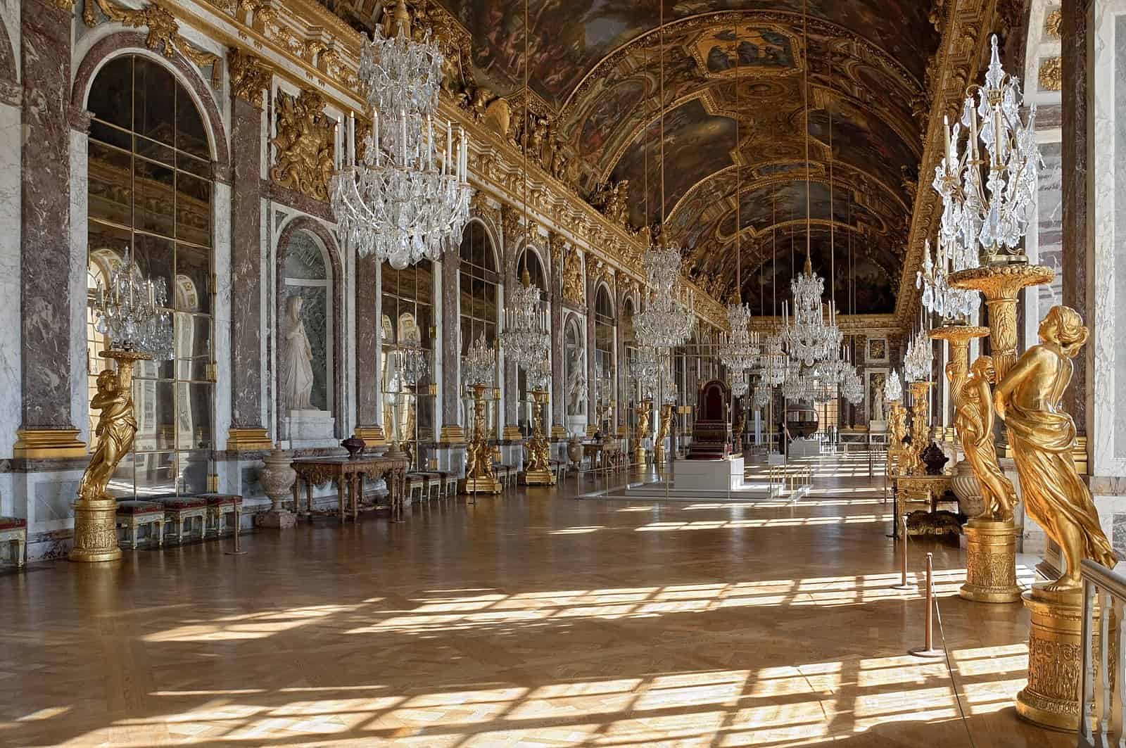 The Hall of Mirrors at the Palace of Versailles, a long gallery of arched mirrors, crystal chandeliers, gilded statues and a painted vaulted ceiling, with sunlight falling across the parquet floor