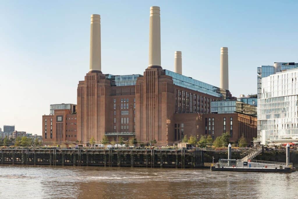 Battersea Power Station seen across the Thames in low sun, all four white fluted chimneys rising from the brick wash towers above the riverside jetty, with glazed rooftop additions and new apartment blocks beside it