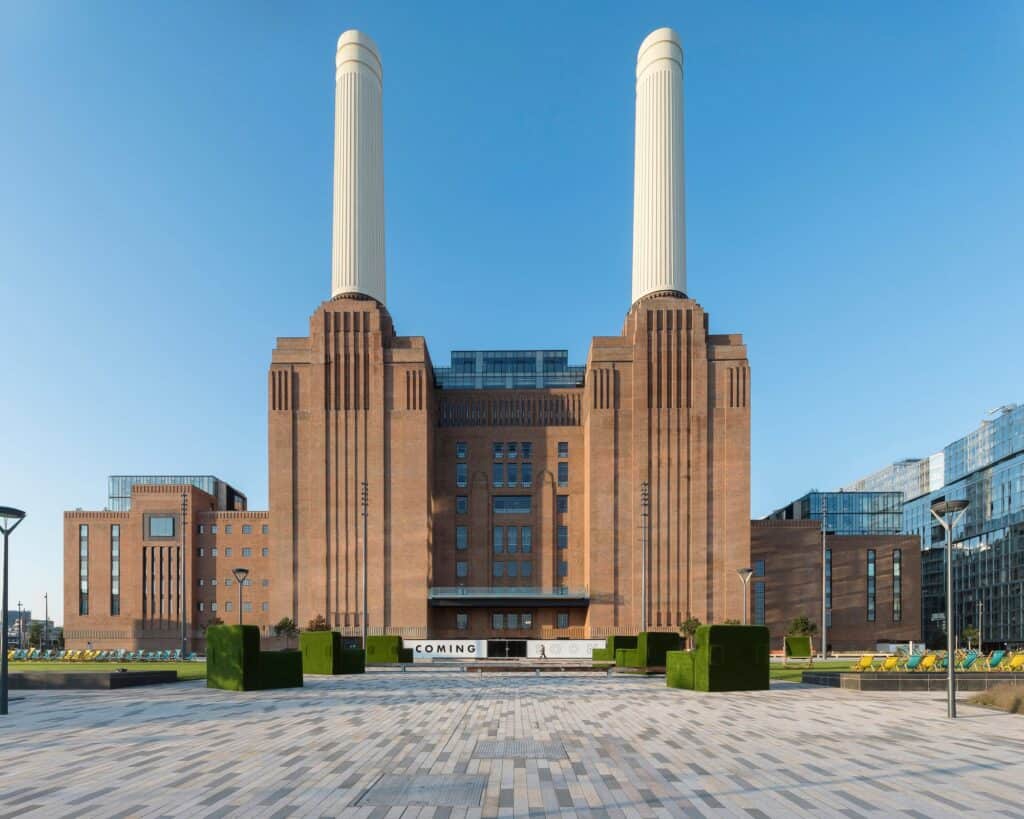 Symmetrical view of Battersea Power Station from the paved plaza, two white chimneys crowning ribbed brick wash towers either side of the central bay, clipped hedges in planters along its base