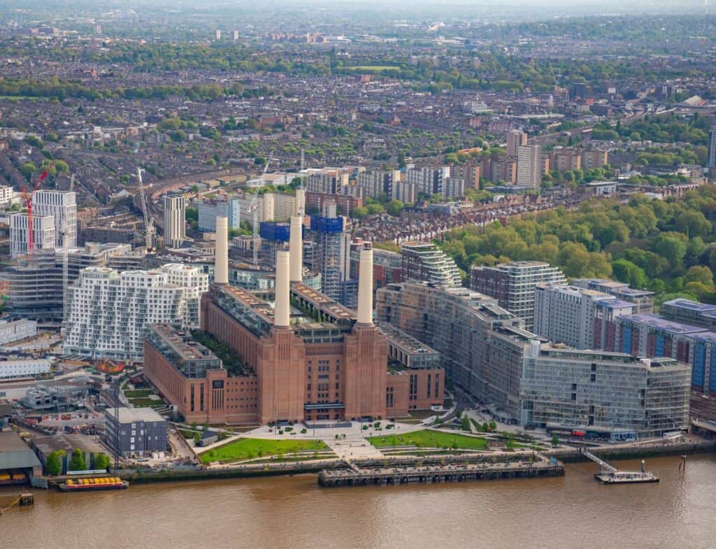 Aerial view of Battersea Power Station on the south bank of the Thames, its four chimneys and brick bulk surrounded by new apartment blocks, riverside lawns and the jetty, with south London stretching to the horizon