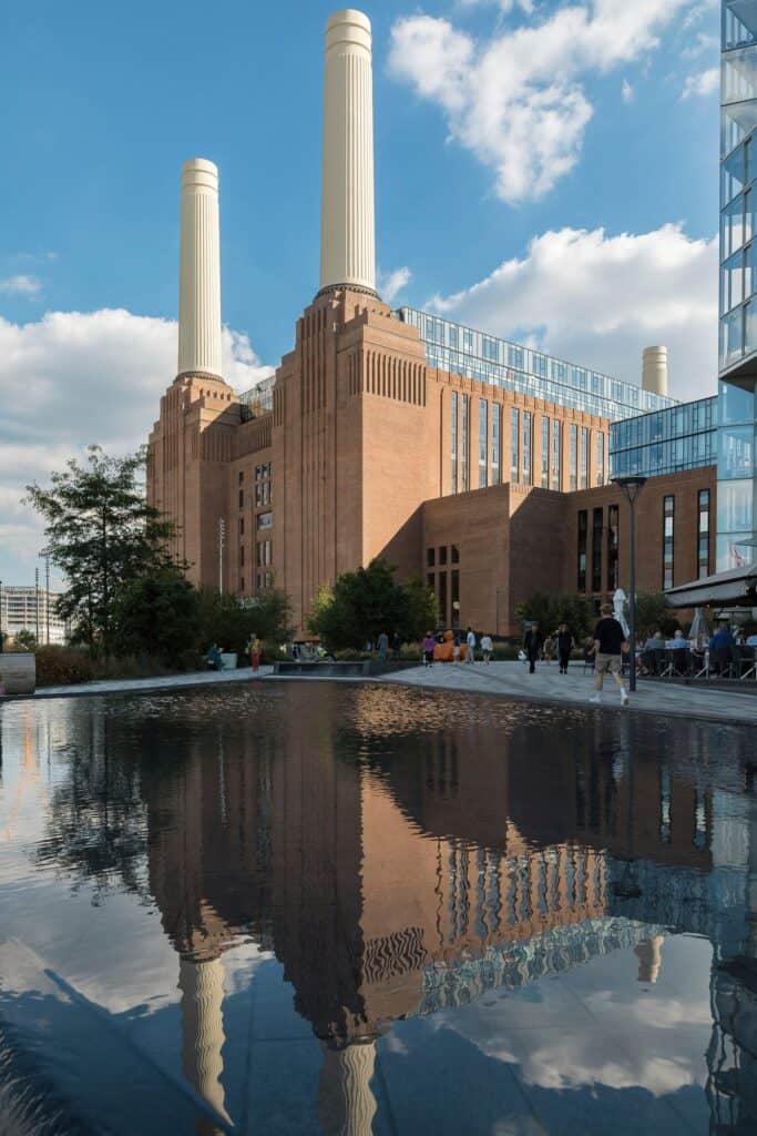Battersea Power Station mirrored in a dark reflecting pool, two chimneys and the brick wash tower rising against white clouds, people walking along the paved terrace beside the water