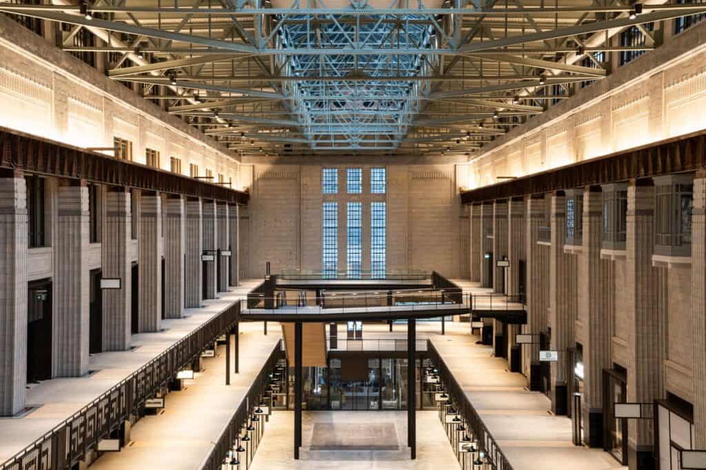 Symmetrical view down Turbine Hall A at Battersea Power Station, tiered shopping galleries between tall faience-clad columns under a glazed steel roof, a bridge crossing the hall towards tall windows at the far end