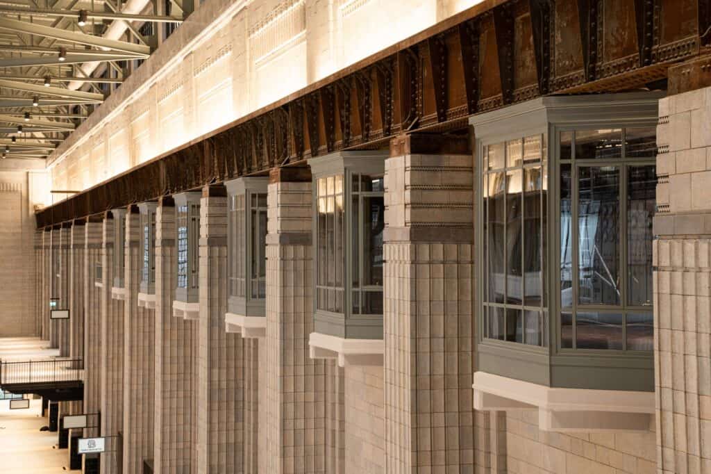 Projecting glazed bay windows set between pale faience-tiled columns in Turbine Hall A at Battersea Power Station, beneath a riveted steel crane rail and warmly lit upper walls