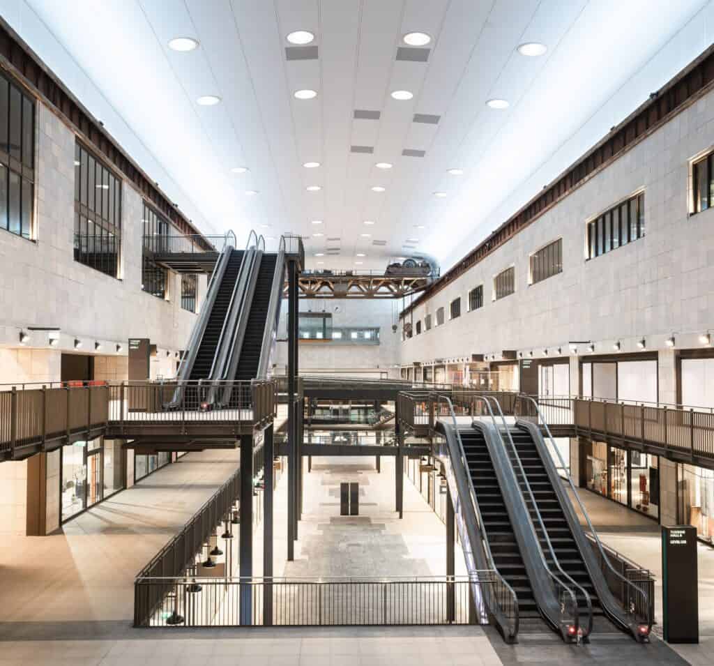 Escalators rising through Turbine Hall B at Battersea Power Station between steel-framed galleries of shops, pale stone walls with rows of tall windows and a white ceiling dotted with round lights