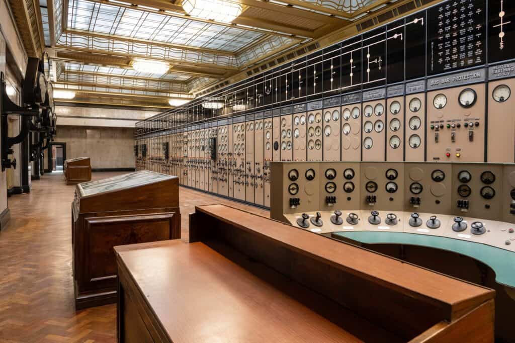 Restored Control Room A at Battersea Power Station with polished timber desks, a curved control desk with rows of dials and switches, long banks of cream switchboards and a herringbone parquet floor beneath a coffered glass ceiling