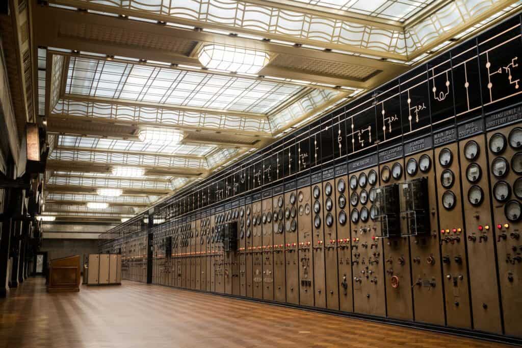 Long bank of restored switchboards with rows of round dials and black mimic diagrams in Control Room A at Battersea Power Station, under a gold-framed coffered glass ceiling above a herringbone parquet floor