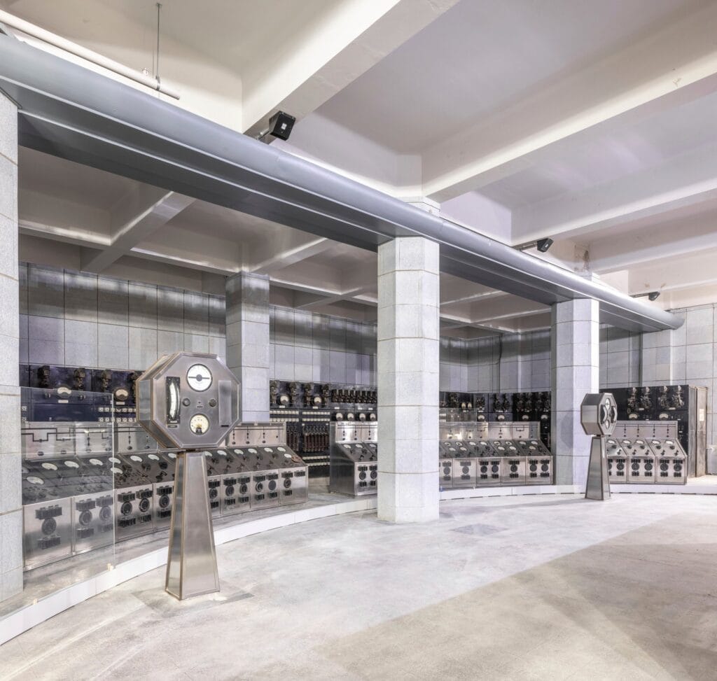 Control Room B at Battersea Power Station, curving banks of stainless steel control panels and two dial-topped instrument stands around a pale terrazzo floor, between square stone-clad columns