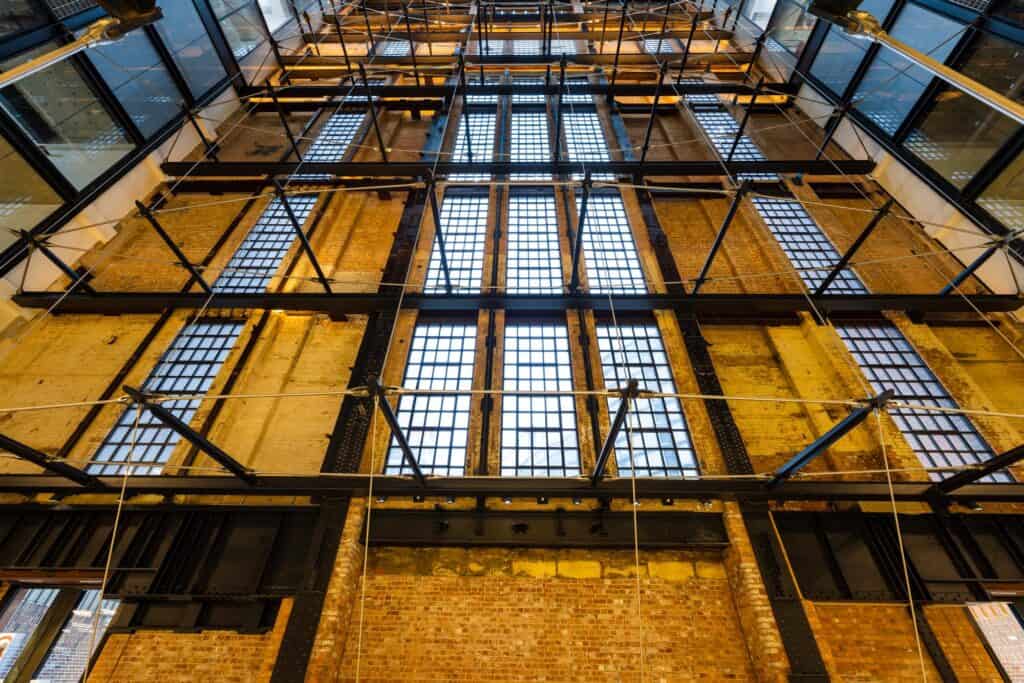Looking up a full-height wall of exposed brick and gridded steel-framed windows inside Battersea Power Station, crossed by black steel bracing and lit by a glazed roof