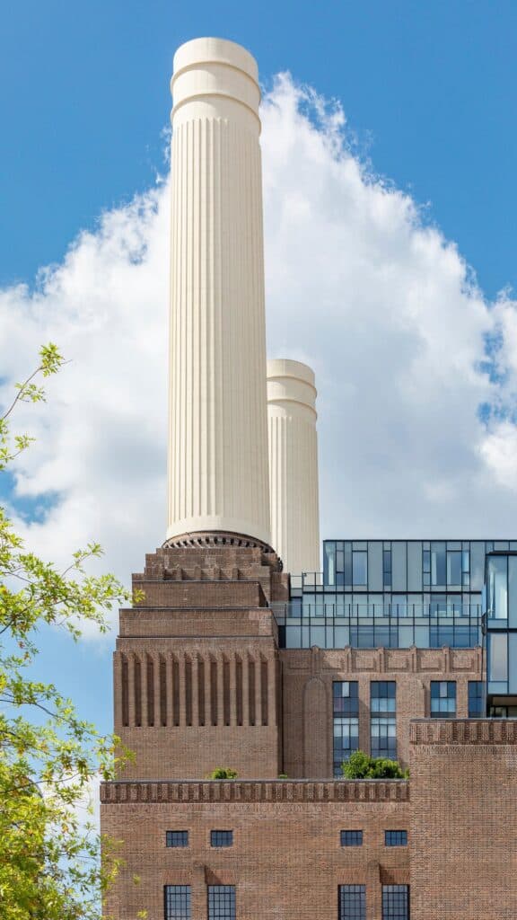 One of the rebuilt fluted white chimneys of Battersea Power Station rising from its stepped brick wash tower against a towering white cloud, glazed rooftop villas behind and green leaves in the foreground