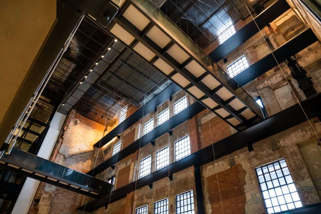Black steel walkways and a staircase bridging a tall void inside Battersea Power Station, past weathered brick walls with patches of old render and rows of gridded windows