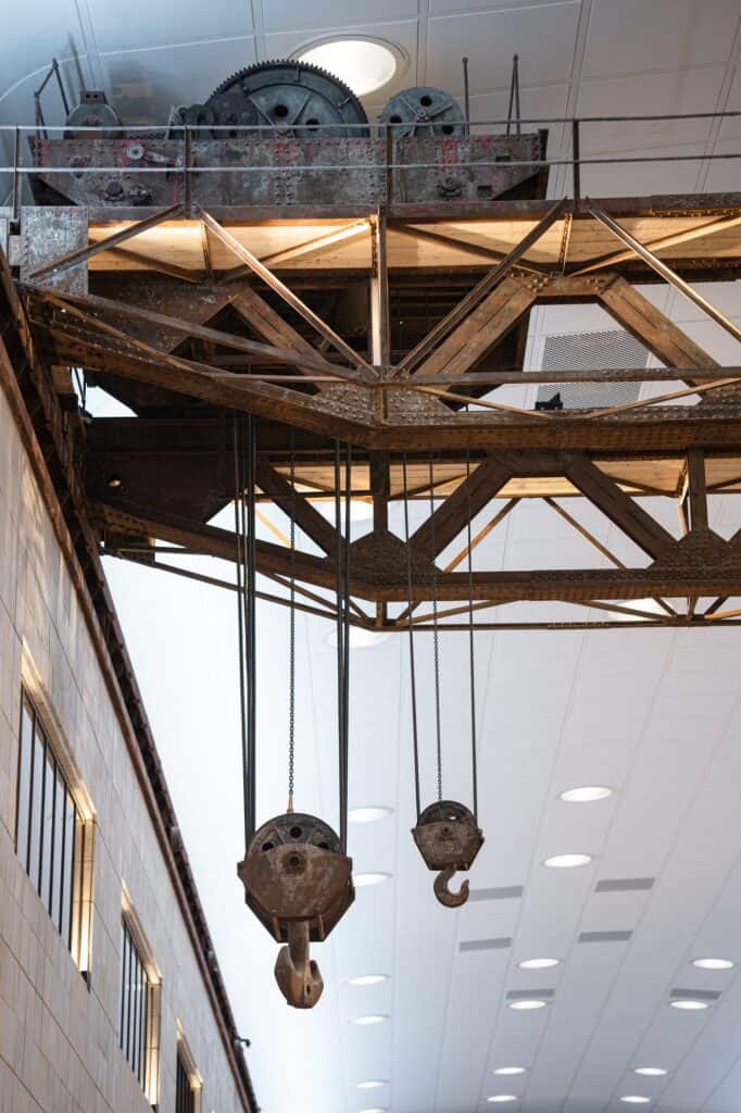 Restored gantry crane with rusted steel trusses, winding gear and two hanging hooks inside a turbine hall at Battersea Power Station, beneath a white ceiling with a round skylight