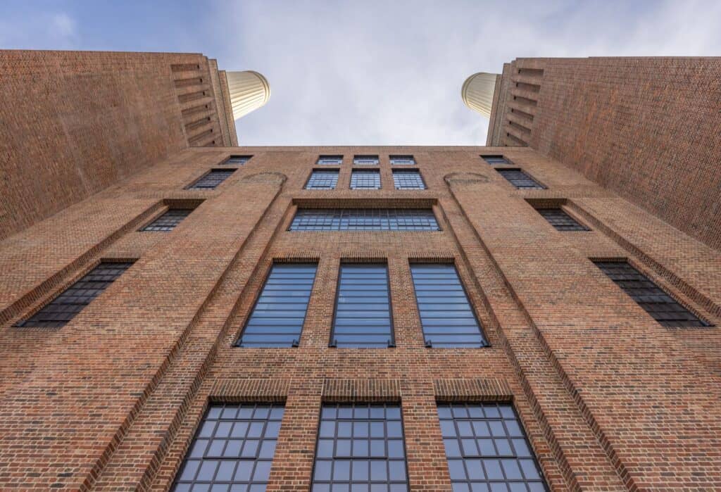 Looking straight up between two brick wash towers of Battersea Power Station to tall steel-framed windows and the bases of the chimneys against a pale sky