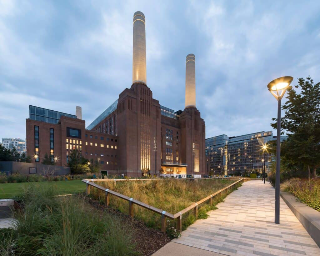 Battersea Power Station at dusk from a planted path in the park, its chimneys uplit against a cloudy sky and windows glowing, a curved street lamp beside the walkway