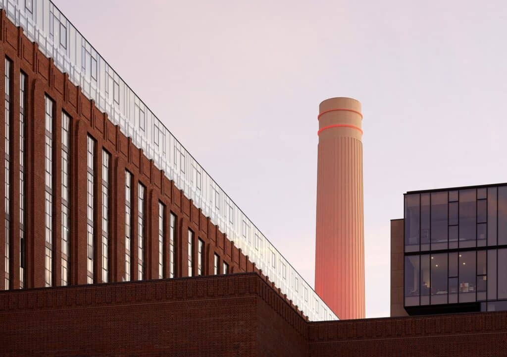 Rebuilt chimney of Battersea Power Station glowing pink at dusk beyond the long brick facade of a Switch House topped with glazed penthouses