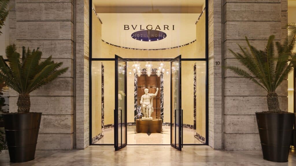 Entrance of Bulgari Hotel Roma between travertine pilasters and potted cycads, glass doors under the BVLGARI name opening onto a marble-lined vestibule with the ancient statue of Augustus