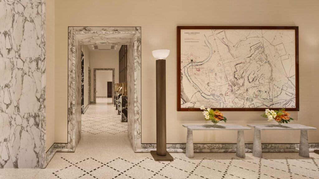Ground-floor corridor at Bulgari Hotel Roma with grey-veined marble door frames, a patterned stone floor, a large framed historic map of Rome over two marble console tables and a bronze floor lamp