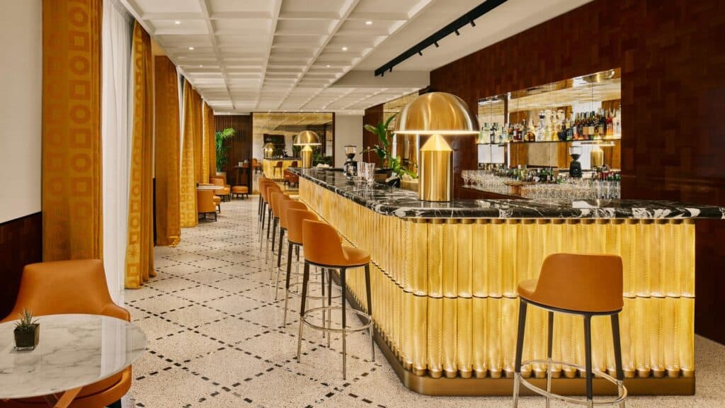 Bulgari Bar at Bulgari Hotel Roma, a long backlit amber glass counter with a black marble top, tan leather bar stools, a gold dome lamp and walnut-panelled shelves of bottles under a coffered ceiling