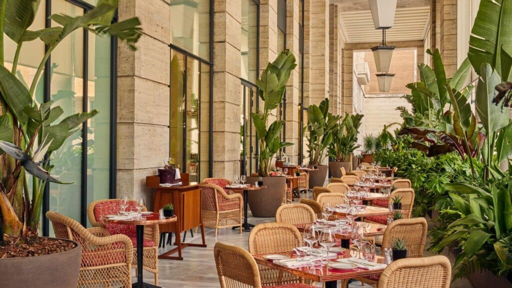 Café under the double-height travertine portico of Bulgari Hotel Roma, rows of woven rattan chairs and red-topped tables between tall glass doors, banana plants and palms
