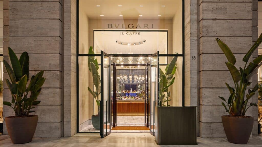 Street entrance to Il Caffè at Bulgari Hotel Roma, glass doors set in a ribbed travertine façade under the BVLGARI Il Caffè sign, flanked by bird of paradise plants in large pots