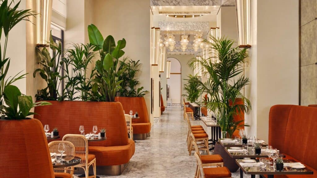 Interior of the café at Bulgari Hotel Roma, rust-orange velvet banquettes and planters with palms lining a marble-floored hall that leads to an arch and a glass chandelier