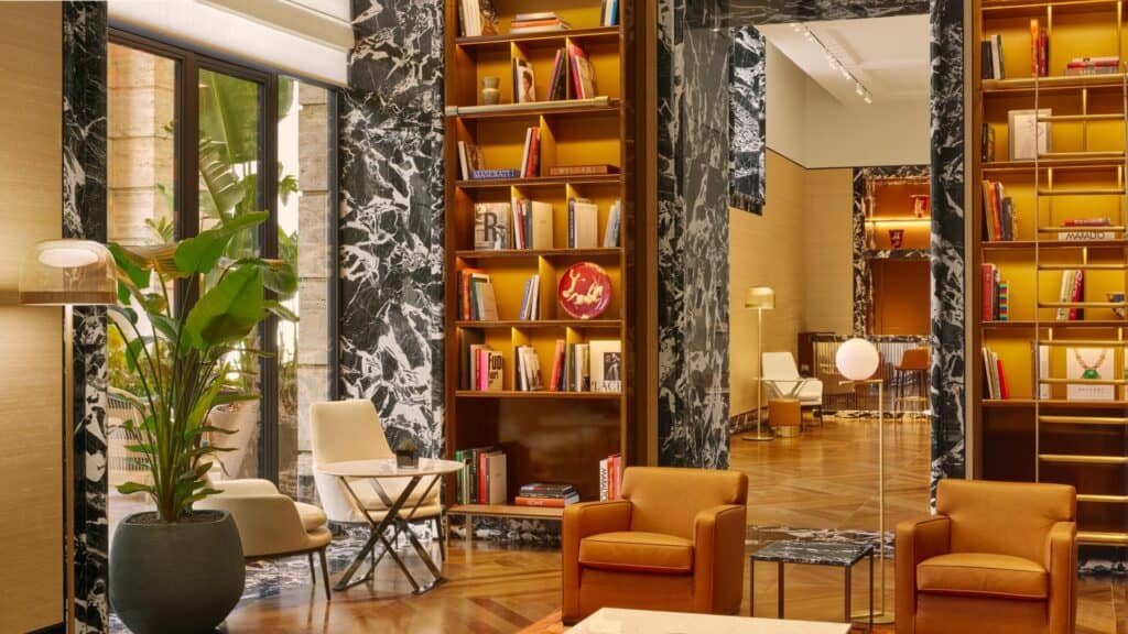 Library at Bulgari Hotel Roma with walnut shelving lit in amber, black and white marble-clad piers, tan leather armchairs, a white lounge chair and a tall plant on a herringbone wood floor