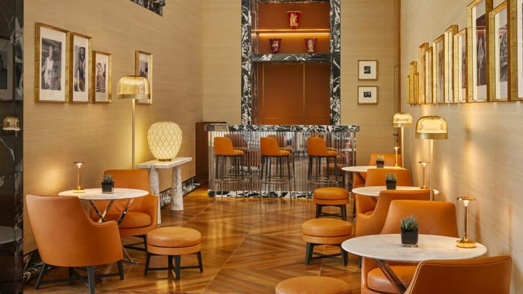 Lounge at Bulgari Hotel Roma with tan leather armchairs and stools around small marble tables on herringbone parquet, framed black and white photographs and a marble-framed bar counter