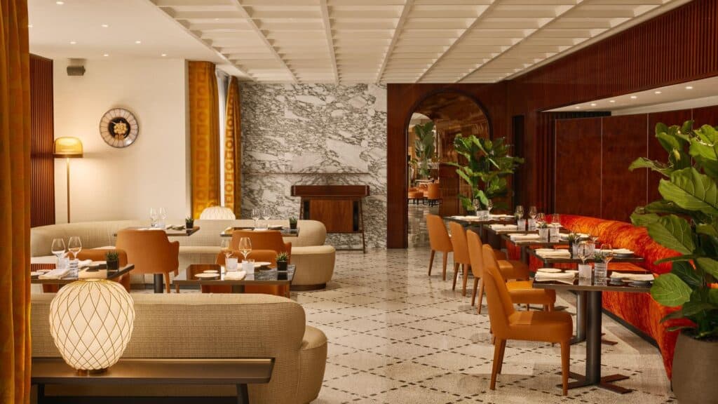 Dining room at Bulgari Hotel Roma beyond beige sofas and a woven globe lamp, with walnut-panelled walls, a grey-veined marble wall, orange leather chairs and a curved red velvet banquette