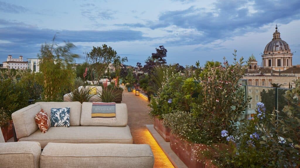 Roof terrace of Bulgari Hotel Roma at dusk, a cream sofa with patterned cushions among planted beds and lavender, with a baroque church dome rising above the Rome rooftops