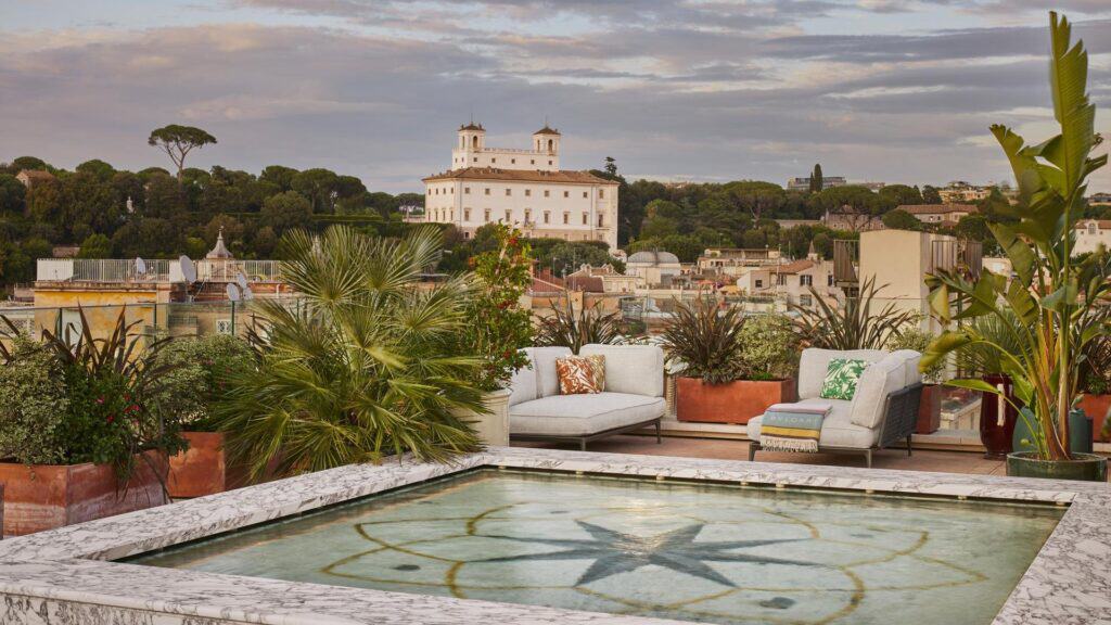 Rooftop garden at Bulgari Hotel Roma around a shallow marble-edged pool with a compass star mosaic, white outdoor sofas and palms, looking towards Villa Medici on the Pincian Hill