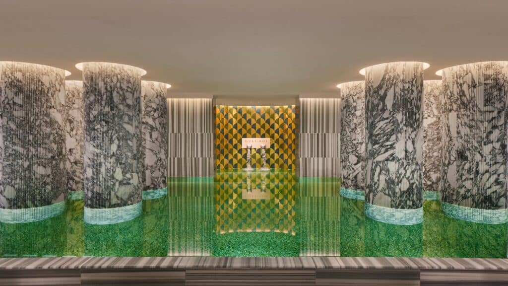 Indoor pool of the Bulgari Spa in Rome, fluted grey and white marble columns standing in green mosaic water before a gold scale-patterned mosaic wall and a lit niche