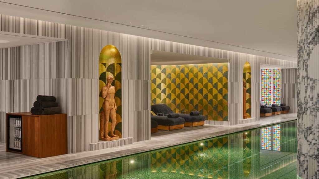 Side of the Bulgari Spa pool in Rome with a gilded classical statue in an arched golden niche, striped marble walls, black loungers in a gold mosaic alcove and a colourful glass window