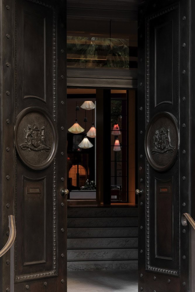 Heavy carved timber entrance doors opening into the lamp-lit interior of Capella Sydney