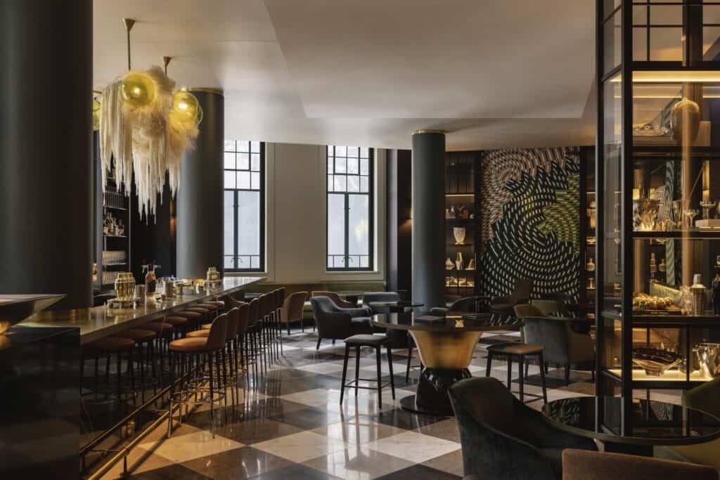 Bar at Capella Sydney beneath a glass chandelier with checkerboard marble floor and steel-framed shelving