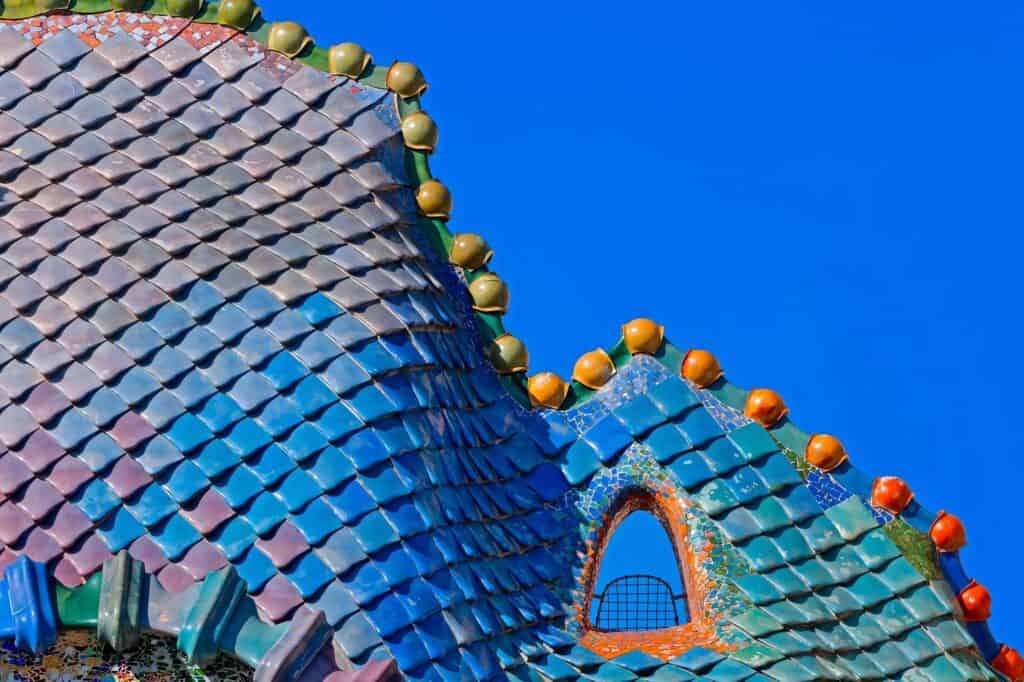 Dragon-back roof of Casa Batlló against a deep blue sky, its scale-like ceramic tiles shading from pink and violet into blue and green, a row of orange ceramic spheres along the ridge and a small arched window