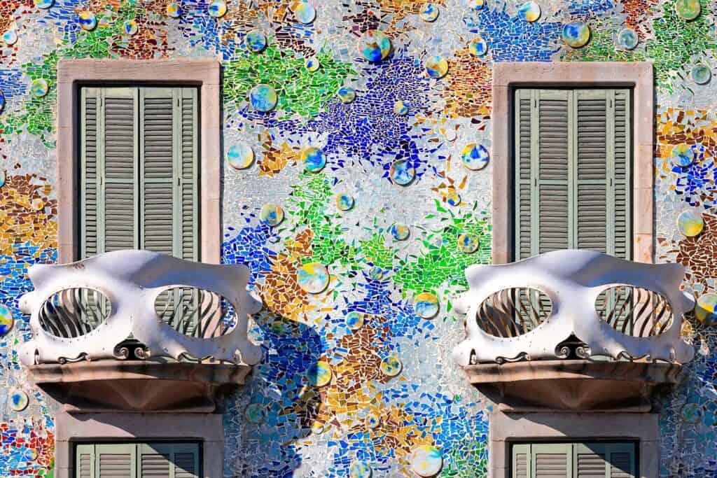 Two white skull-like balconies on the facade of Casa Batlló against shimmering trencadís of blue, green and gold glass and ceramic discs, with grey wooden shutters behind